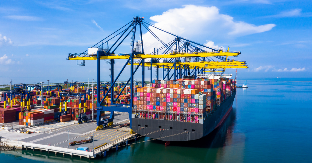 A large container ship loaded with brightly colored containers docked at a port, alongside massive yellow gantry cranes and stacks of shipping containers.